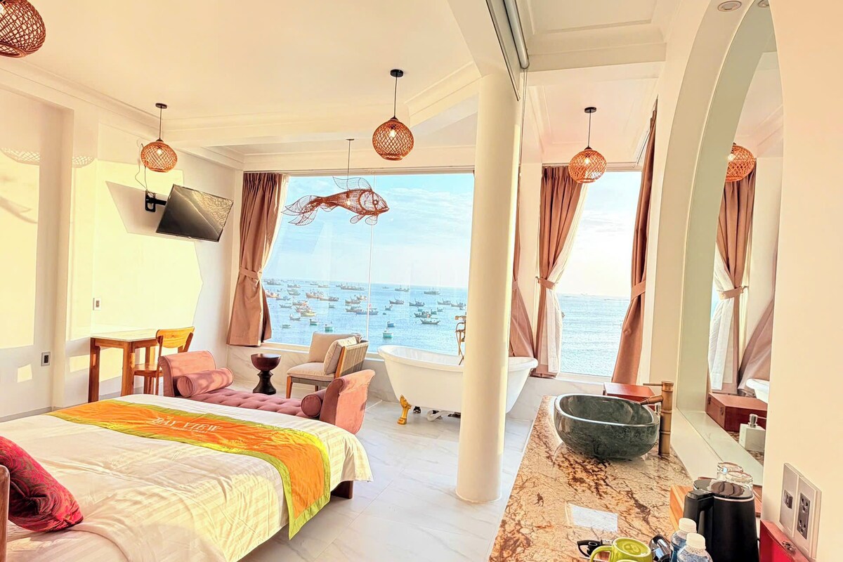 A hotel room with a bed, a desk, a bathtub, and a view of the sea.
