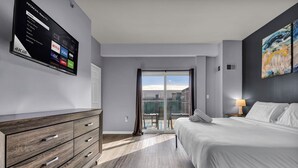 Habitación de hotel moderna con un televisor de pantalla plana grande montado en la pared, cama con ropa de cama blanca, cómoda de madera y vista a la ciudad a través de una puerta corrediza de vidrio.