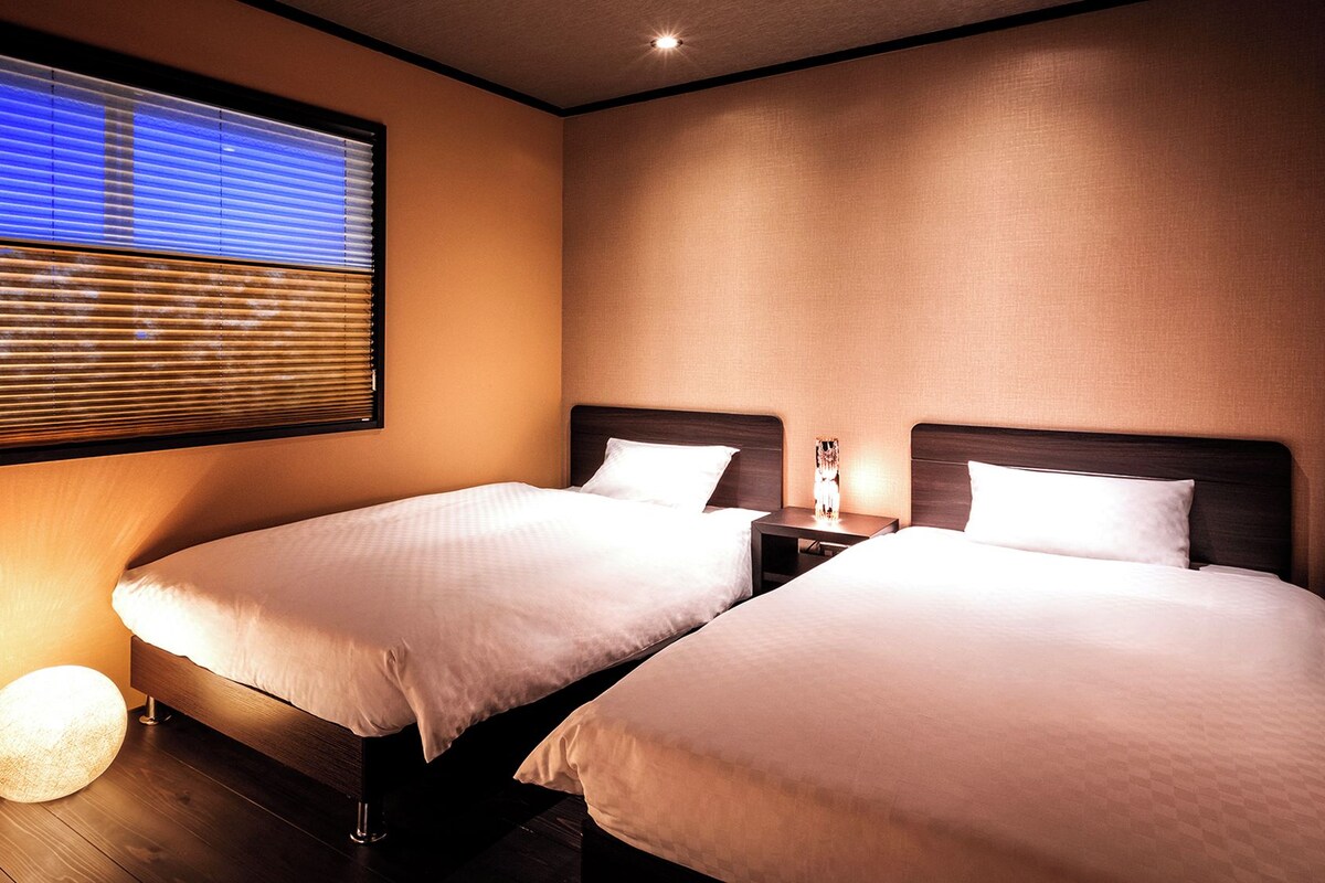 Een hotelkamer met twee bedden, een nachtkastje, een lamp en een raam met jaloezieën.