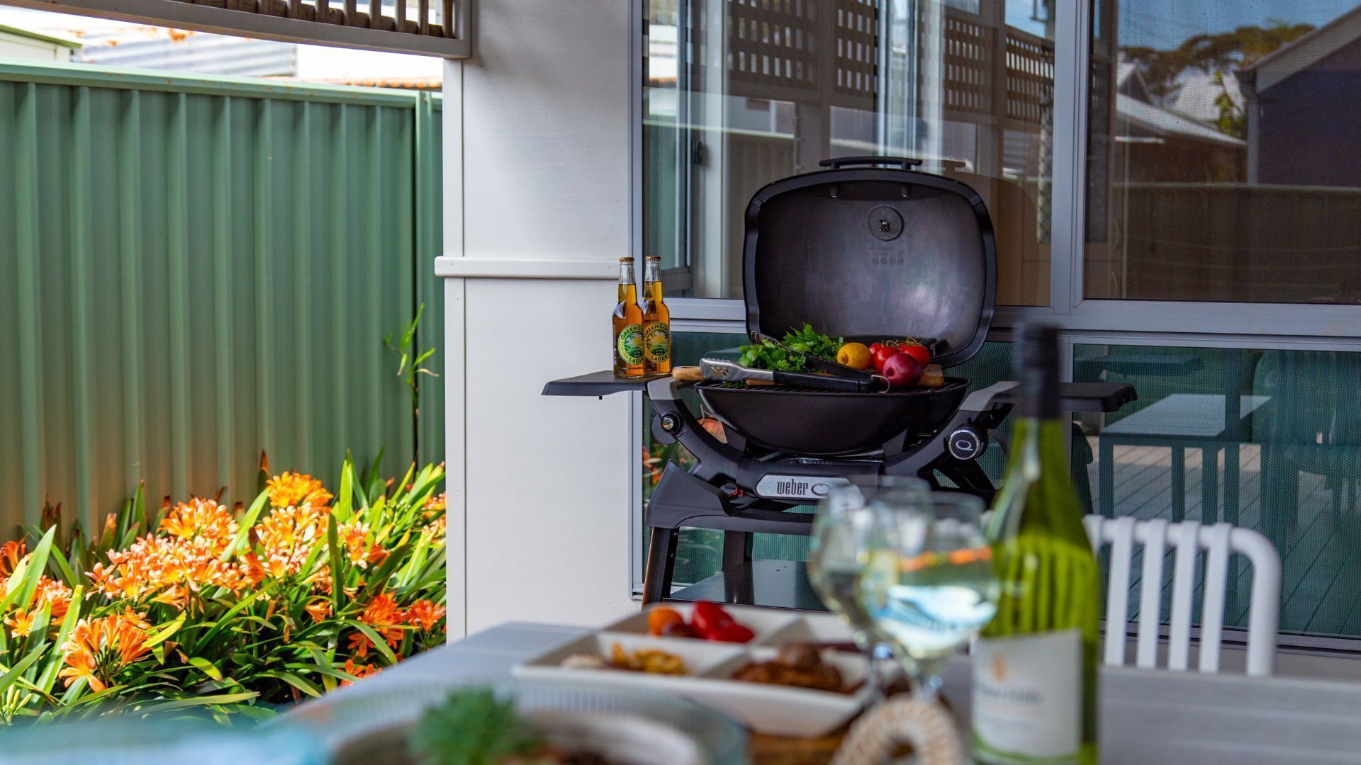 A patio with a Weber grill, a table with food, a bottle of wine, and orange flowers.
