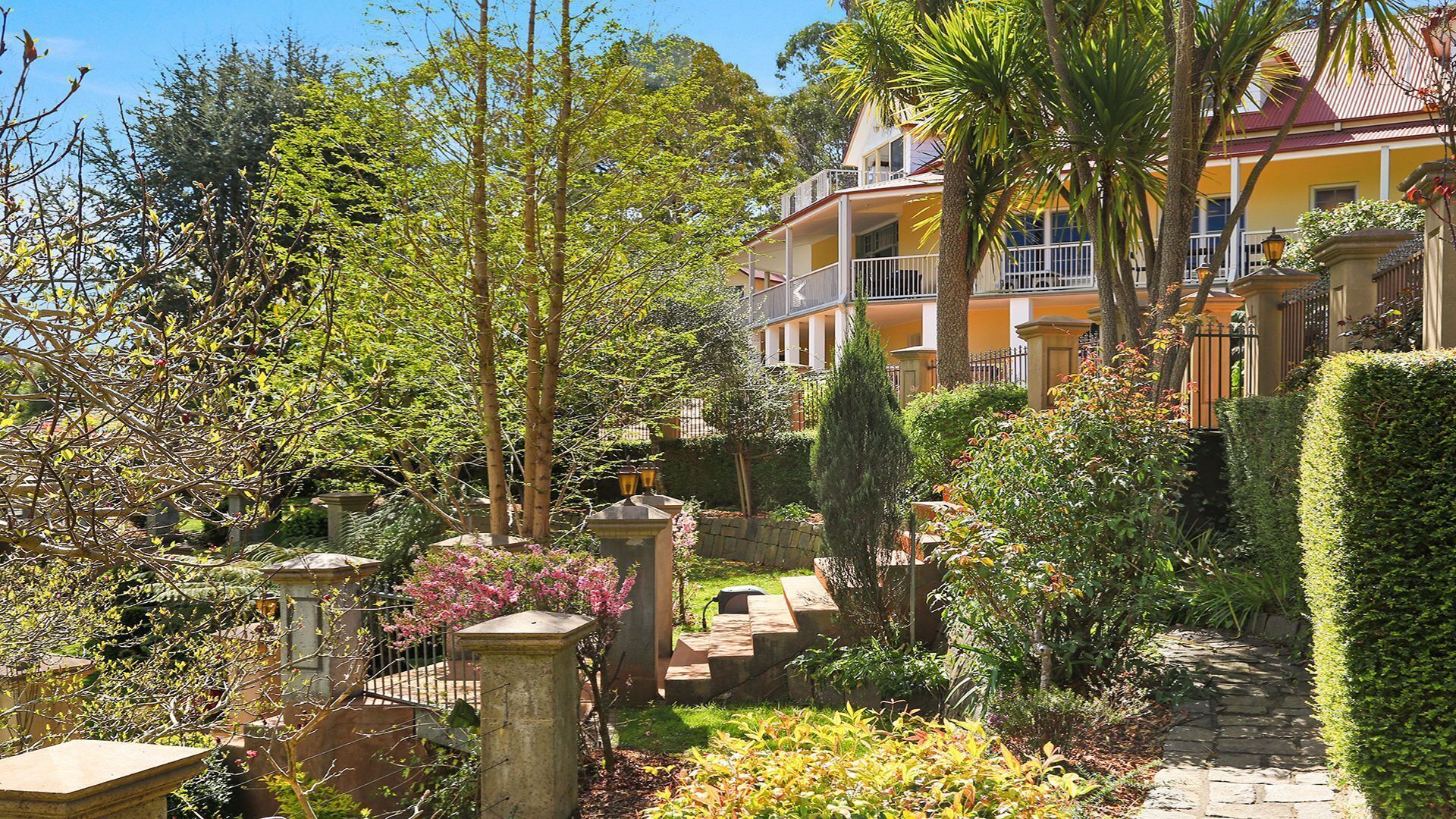 A garden with a stone pathway, a fountain, and a yellow building with a balcony.
