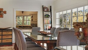 A dining area with a glass table and patterned chairs, a bookshelf with books and decorative items, and a view of the outdoors through large windows. - Southern Grace - the perfect Southern Highlands escape, spacious, stylish and serene (Bowral)