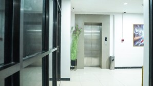 Een schone, moderne lobby met een glazen deur, een potplant, een lift en een ingelijst schilderij aan de muur.