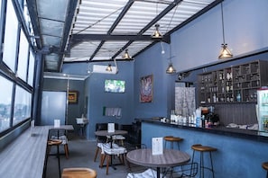 Zona de bar moderna con techos altos, vigas a la vista e iluminación colgante. Hay taburetes de bar, mesas y una barra con una exhibición de bebidas.