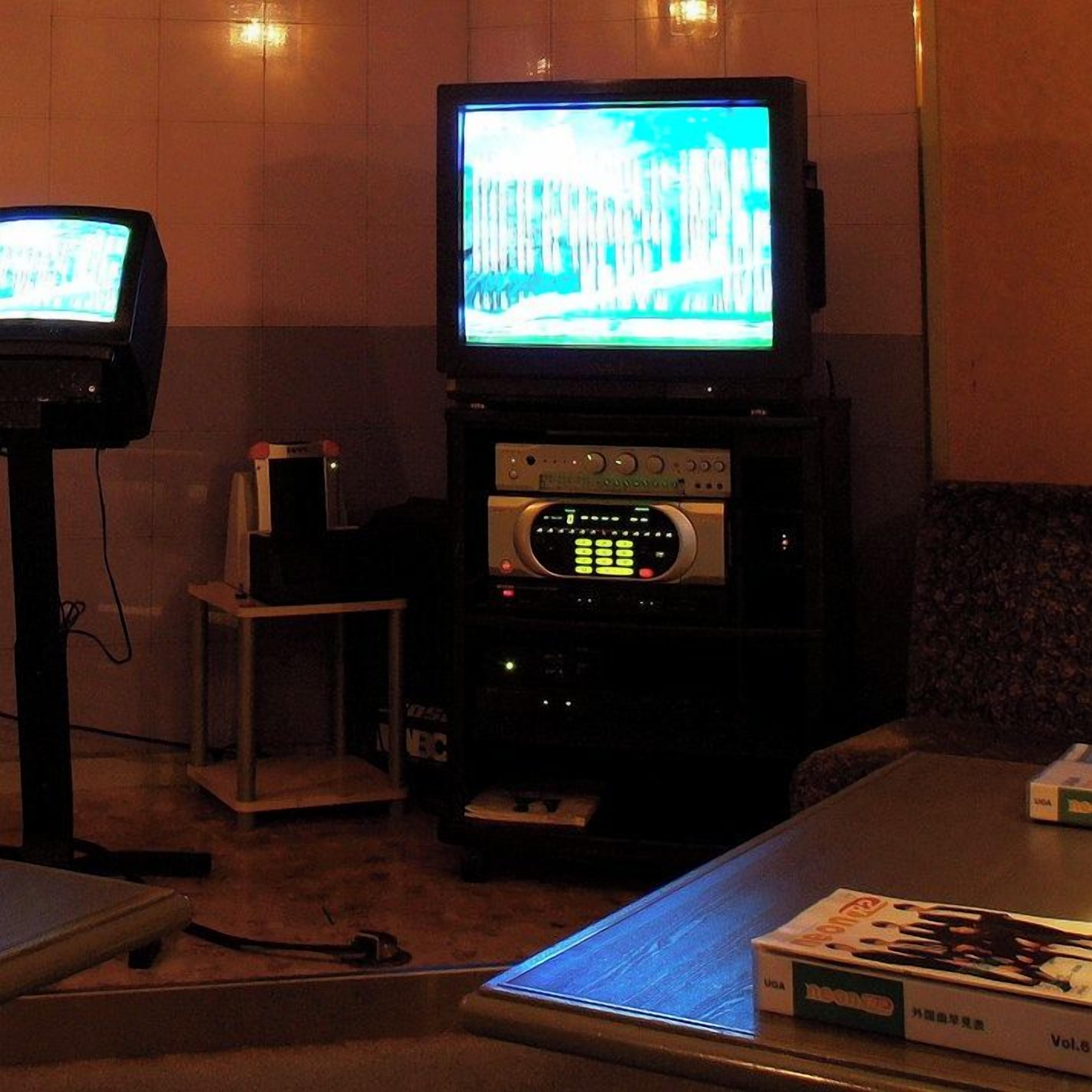 Una habitación con una televisión sobre un mueble, una pequeña mesa con una caja y un altavoz en un estante.