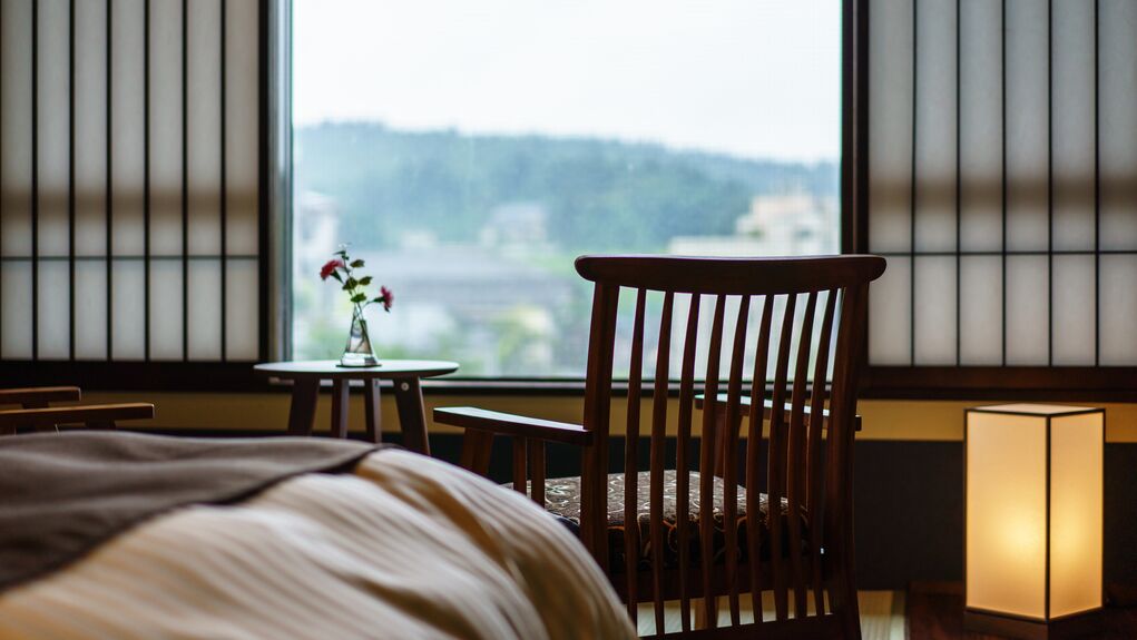 A hotel room with a bed, a wooden chair, a small table with a vase, and a window with a view of a cityscape.