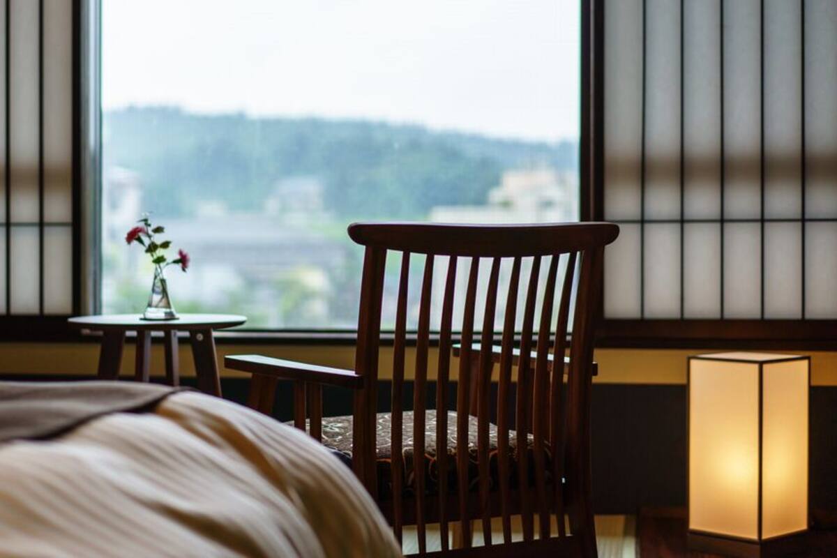 A hotel room with a bed, a wooden chair, a small table with a vase, and a window with a view of a cityscape.