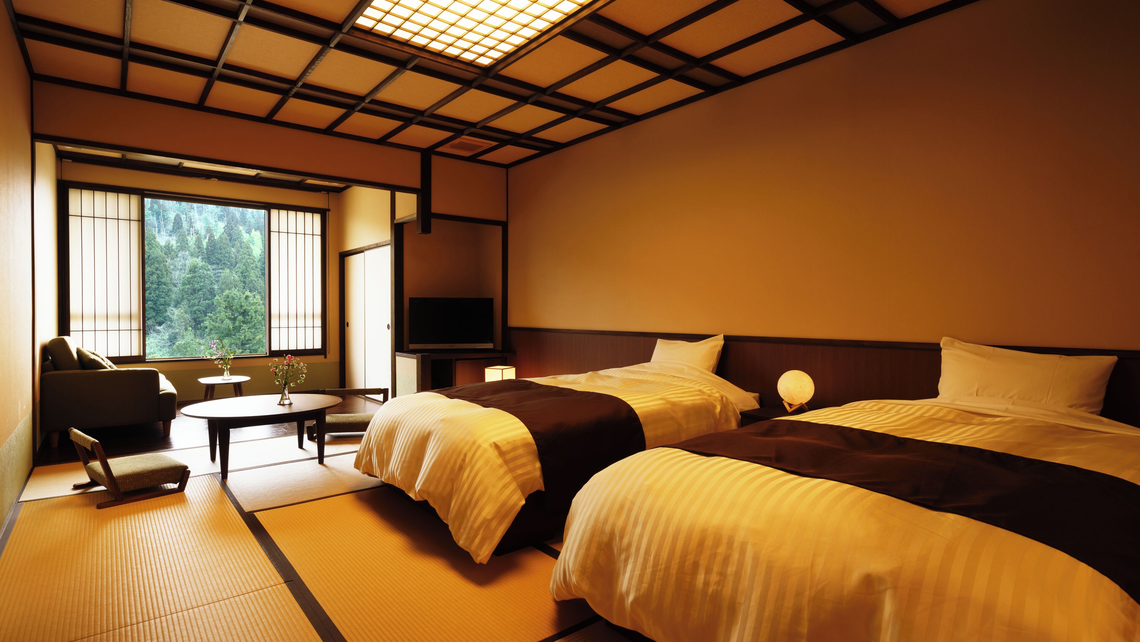 Quarto de hotel com duas camas, televisão, uma mesinha com um vaso e vista para árvores pela janela.