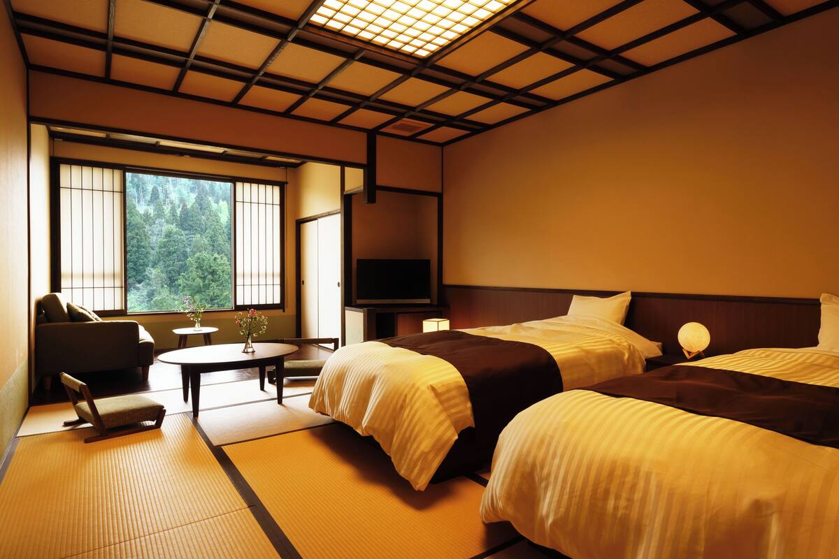 A hotel room with two beds, a TV, a small table with a vase, and a view of trees through the window.
