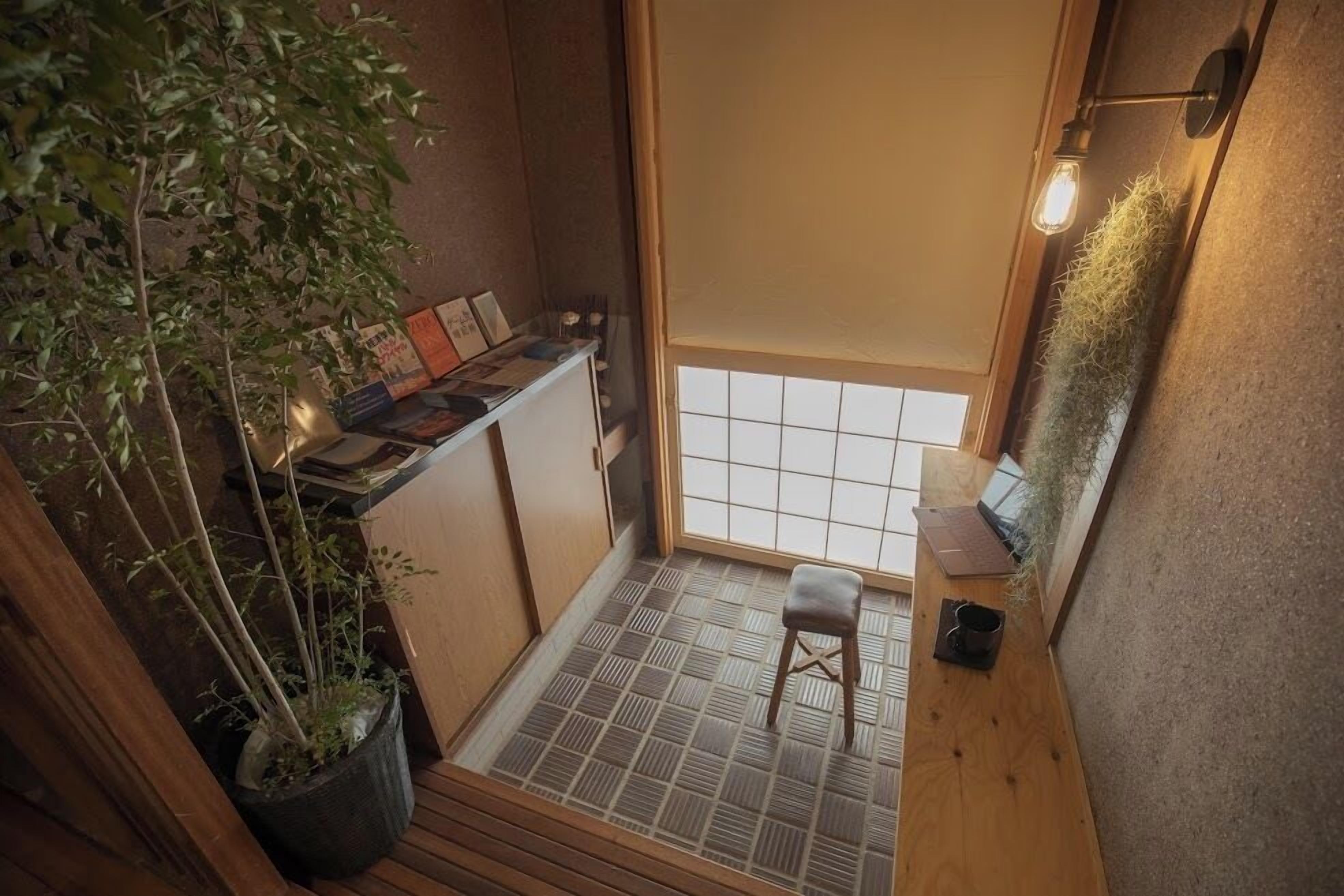 Une petite pièce avec un sol recouvert de tatamis, une basse table en bois, un tabouret, une étagère remplie de livres et une plante en pot.