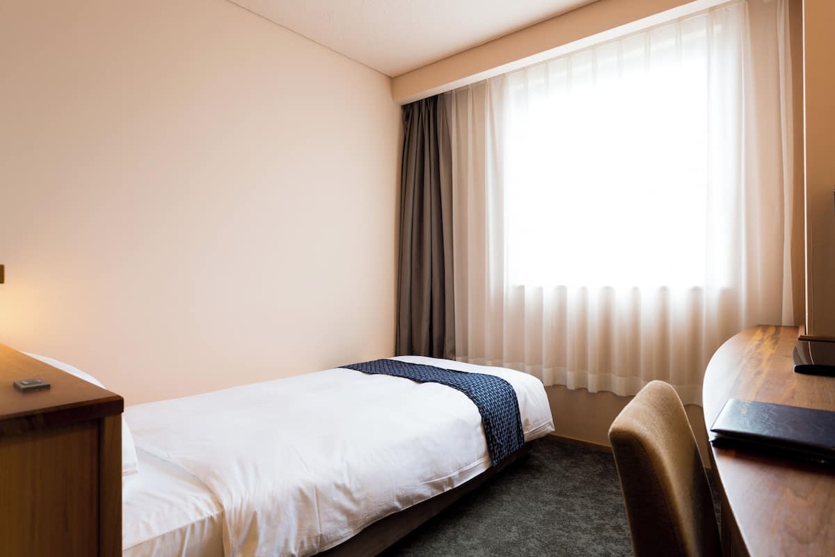 A hotel room with a single bed, a desk, a chair, and a window with curtains.