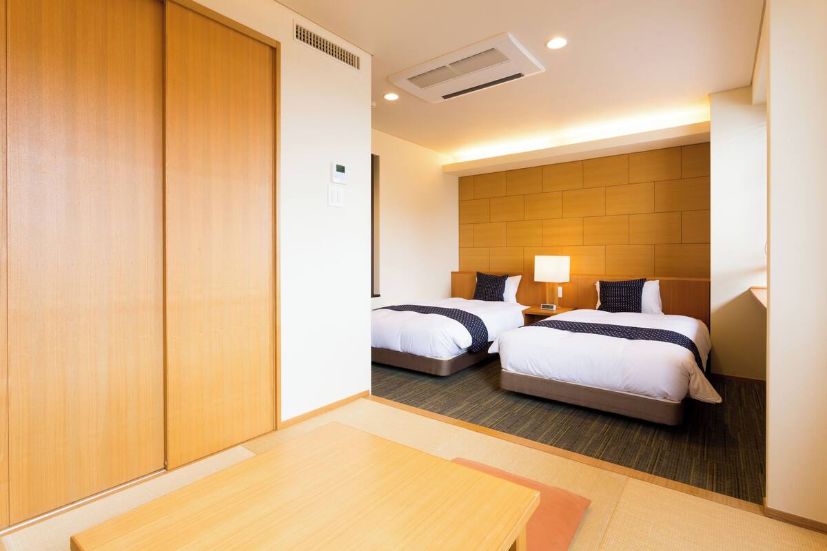 A hotel room with two beds, a wooden wardrobe, a table, and a lamp.