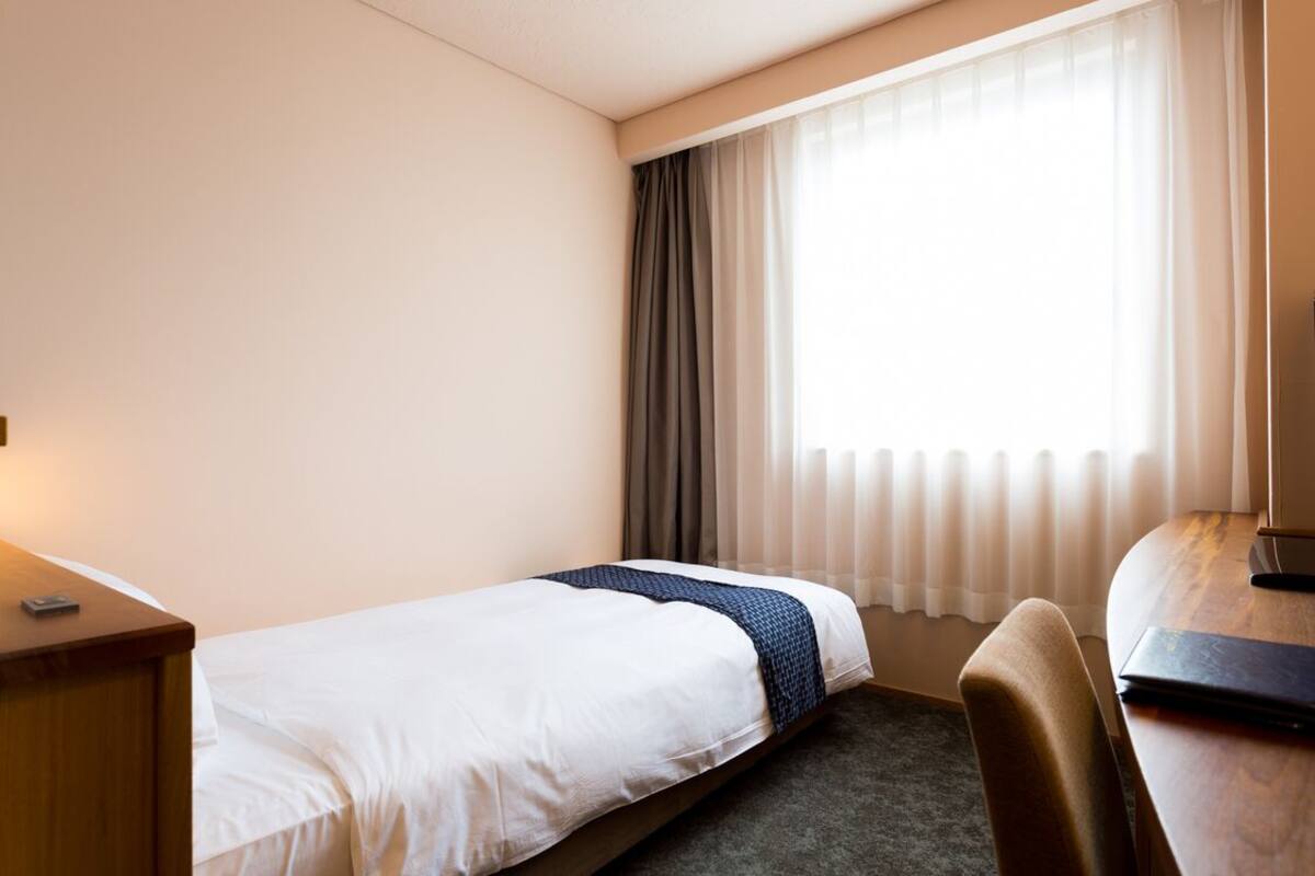 A hotel room with a single bed, a desk, a chair, and a window with curtains.