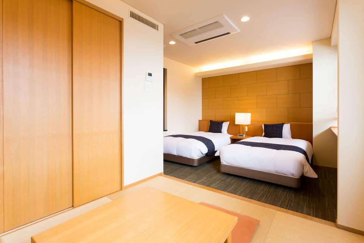 A hotel room with two beds, a wooden wardrobe, a table, and a lamp.