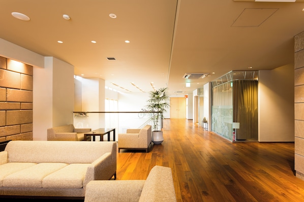 A modern hotel lobby with a wooden floor, seating area with sofas, a reception desk, and a large potted plant. - Takanoko Hotel (Matsuyama)