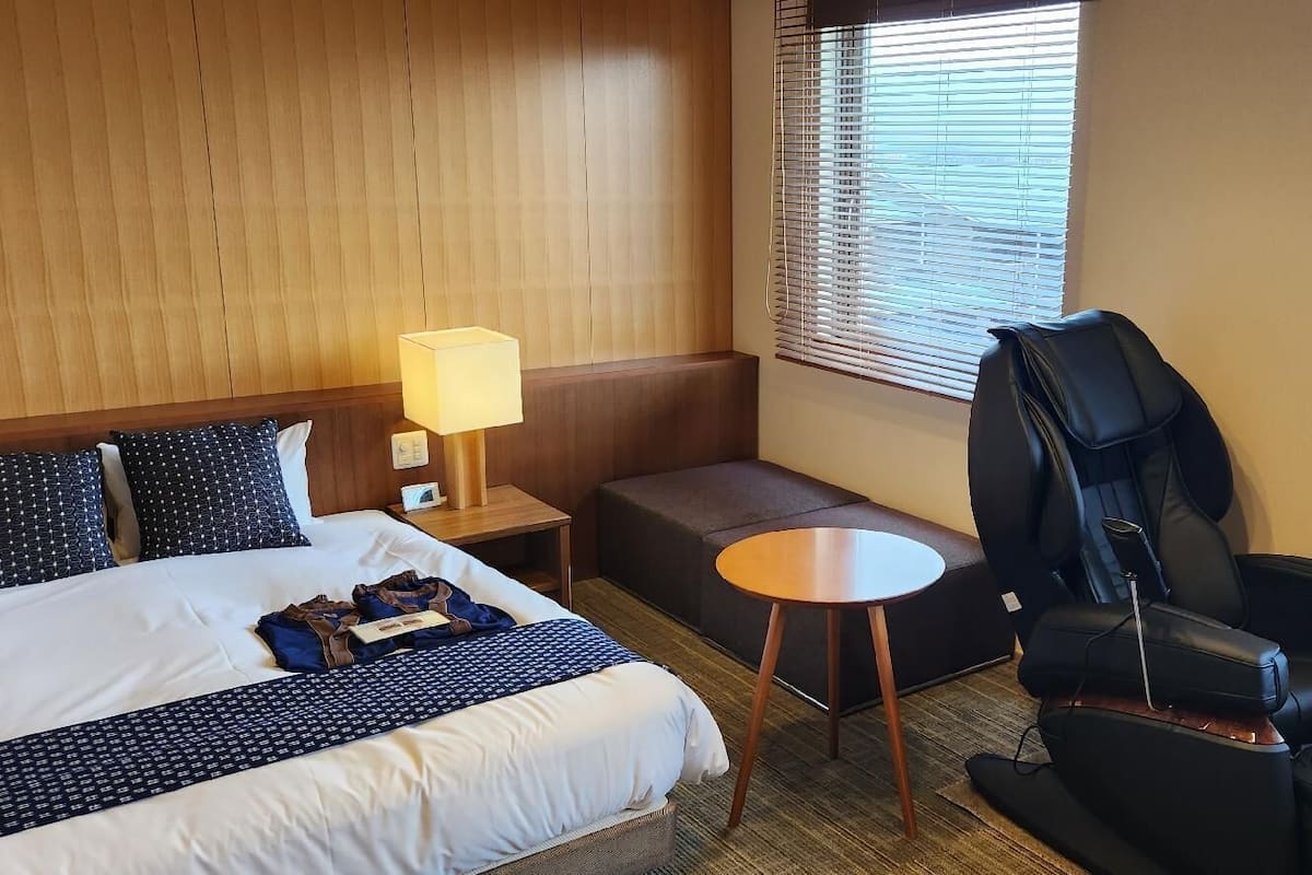 A hotel room with a bed, a desk, a massage chair, and a window with blinds.
