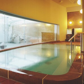 A swimming pool with a glass wall, a wooden deck, and a stone wall in the background. - Takanoko Hotel (Matsuyama)