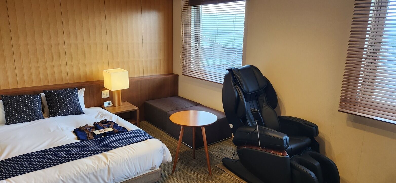 A hotel room with a bed, a desk, a massage chair, and a window with blinds.