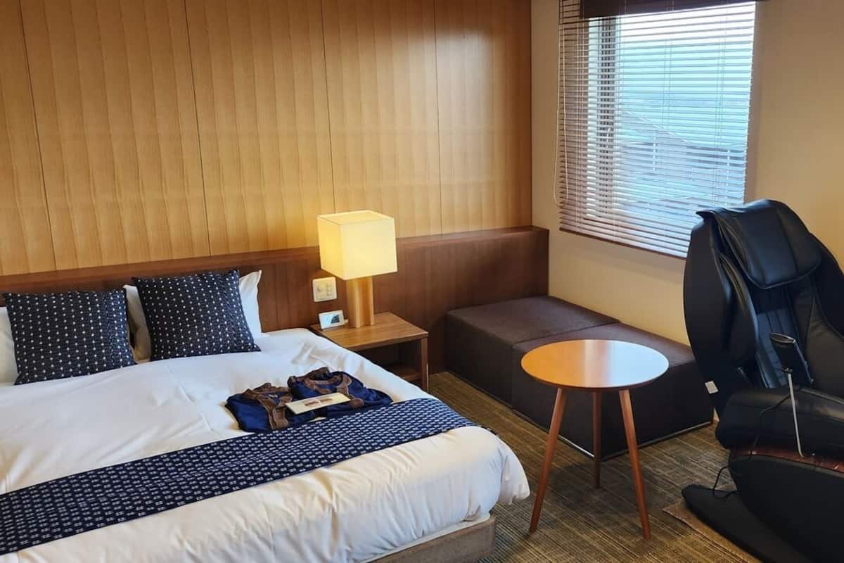 A hotel room with a bed, a desk, a massage chair, and a window with blinds.
