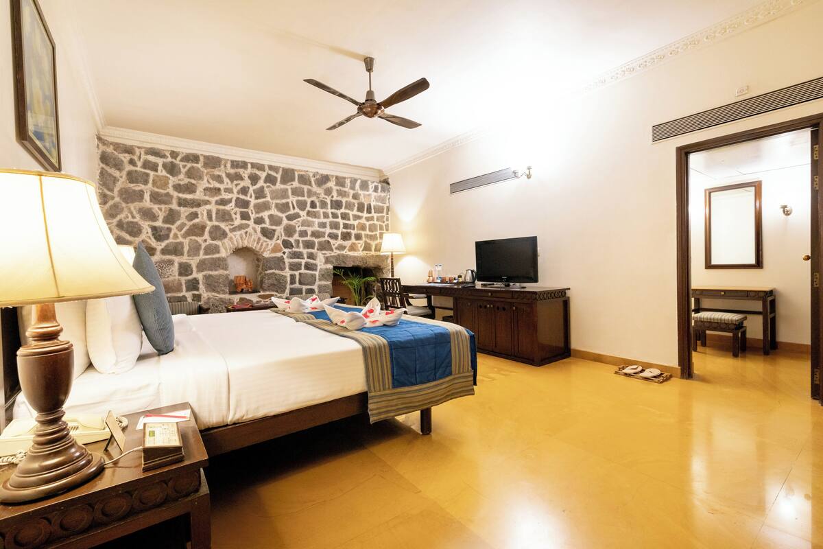 A hotel room with a stone fireplace, a bed with a blue blanket, a television on a wooden cabinet, and a ceiling fan.