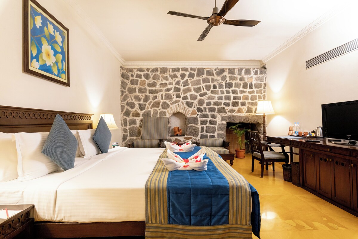 A hotel room with a stone wall, a bed with white linens, a blue bedspread, a wooden headboard, a ceiling fan, a television, and a desk with a lamp.