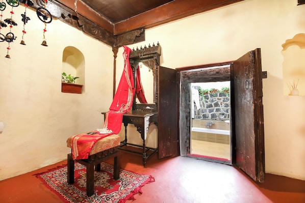 Une pièce comprenant une chaise en bois, une petite table, un miroir et un tissu rouge. Il y a une fenêtre donnant sur un mur de pierre et un jardin.