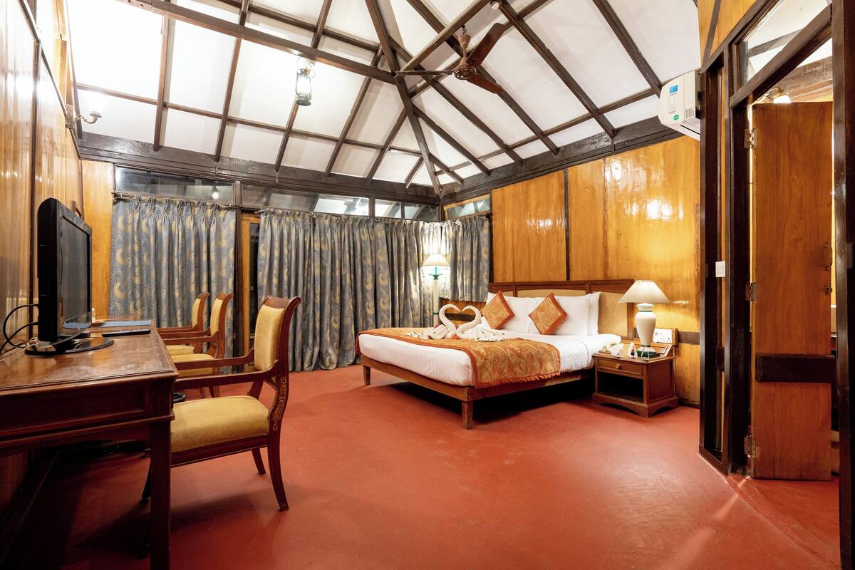 A hotel room with a wooden ceiling, a bed with a heart-shaped pillow, a desk with a chair, and a nightstand with a lamp.
