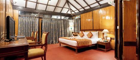 A hotel room with a wooden ceiling, a bed with a heart-shaped pillow, a desk with a chair, and a nightstand with a lamp.