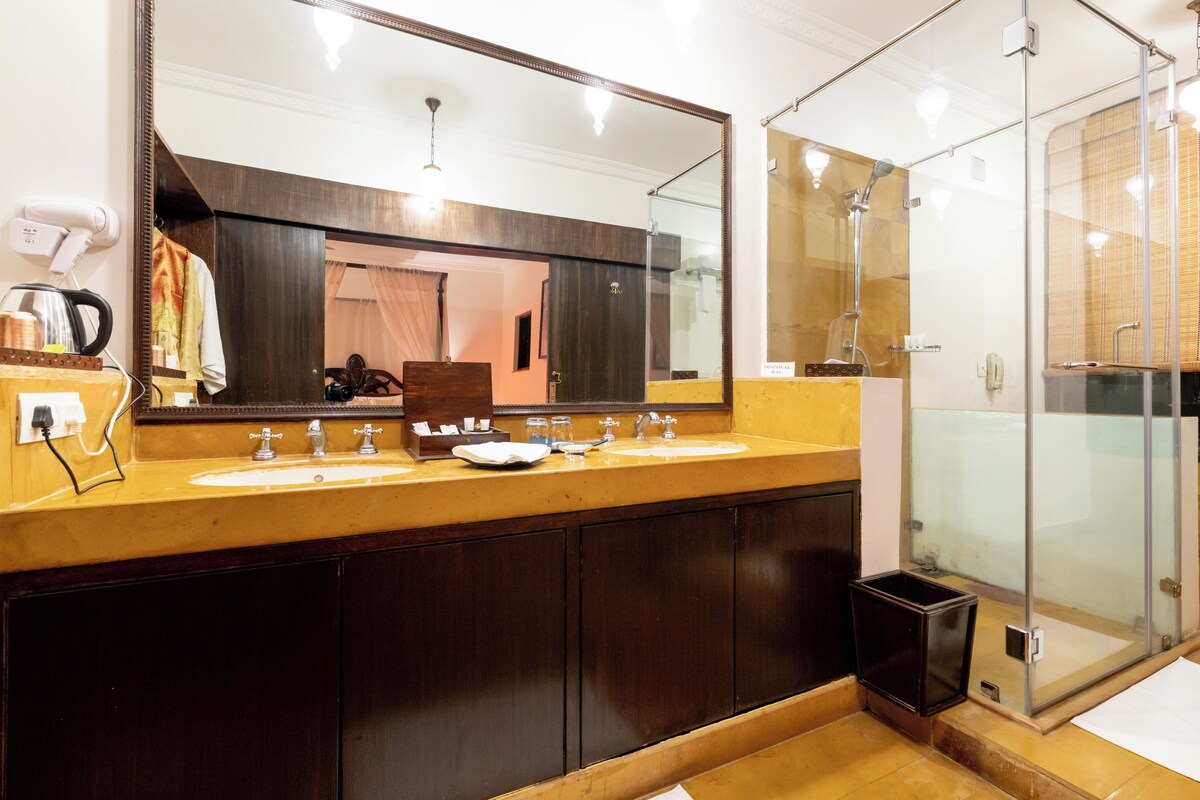 A bathroom with a double sink, a large mirror, a glass shower enclosure, and a trash bin.