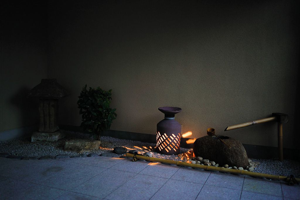Un jardín japonés tradicional con un farol de piedra, un jarrón grande y un rastrillo de bambú.