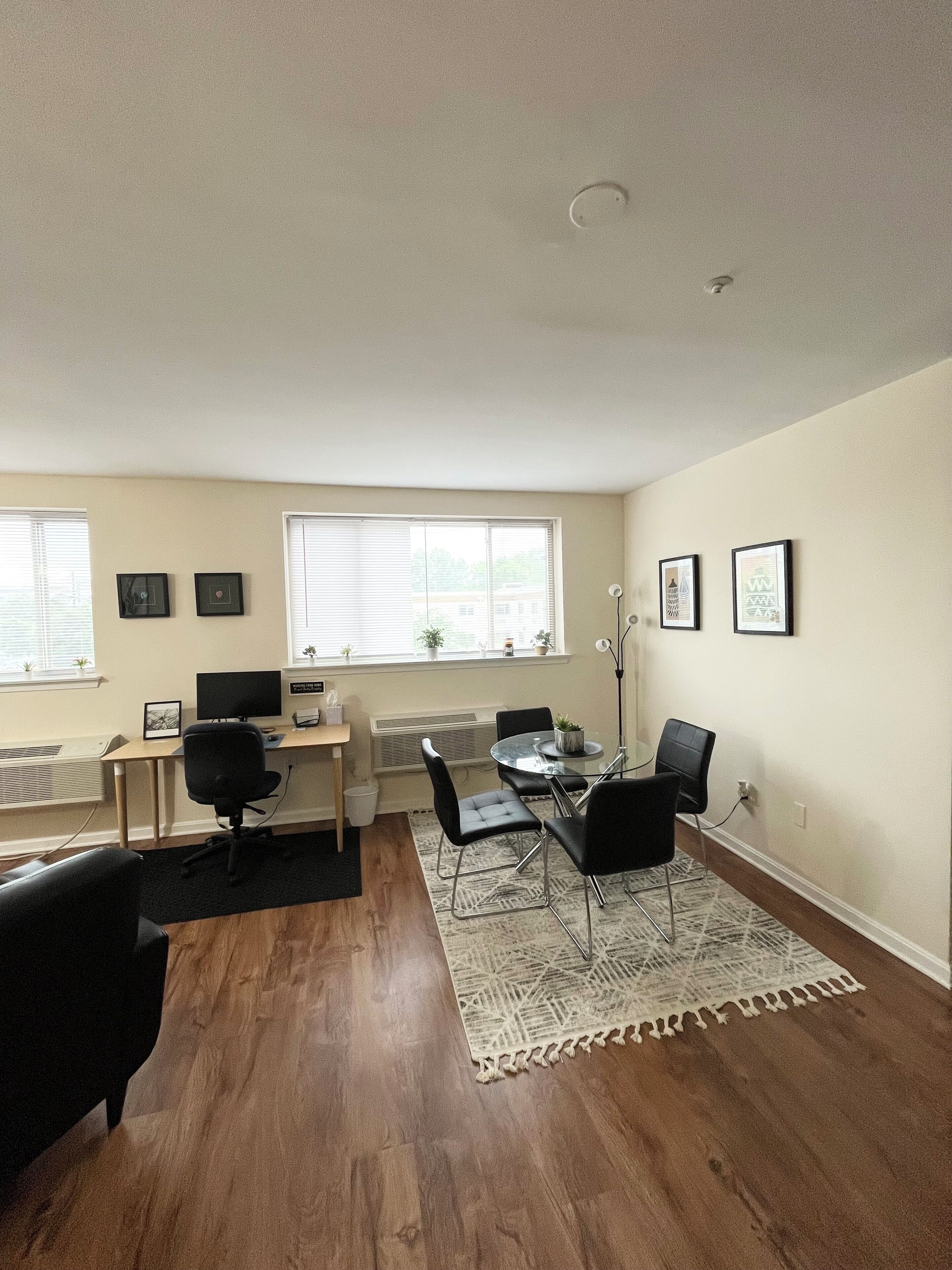 A modern living room with a wooden floor, a dining table with chairs, a computer desk, and a large window with blinds.
