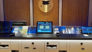 Un mostrador de recepción con una computadora portátil mostrando un menú digital, una placa dorada con texto japonés y un panel de pared de madera.