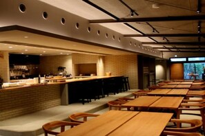 A restaurant with long wooden tables and chairs, a brick counter with a bar area, and a view of the outdoors through large windows. - Arita Huis (Arita)