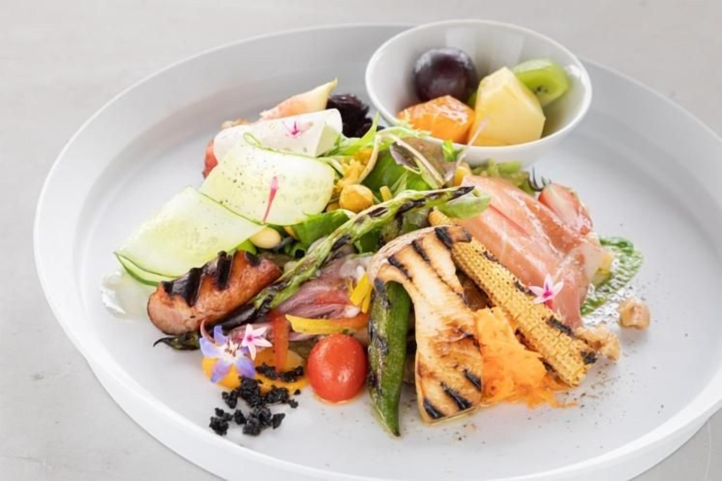 Ein Teller mit verschiedenen gegrillten Gemüsesorten und Fleisch, dazu eine kleine Schale Obst.