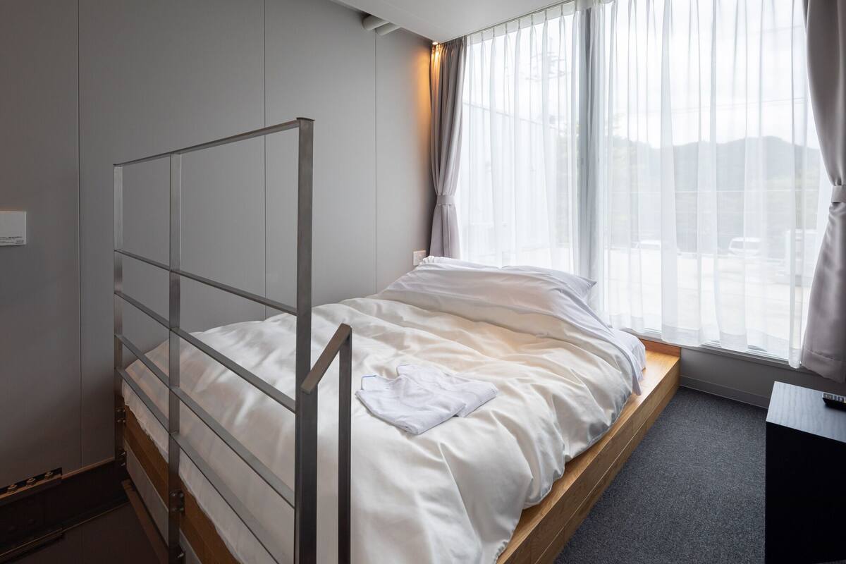 Uma cama de solteiro com estrutura metálica, uma janela com cortinas leves e uma mesa com um controle remoto.
