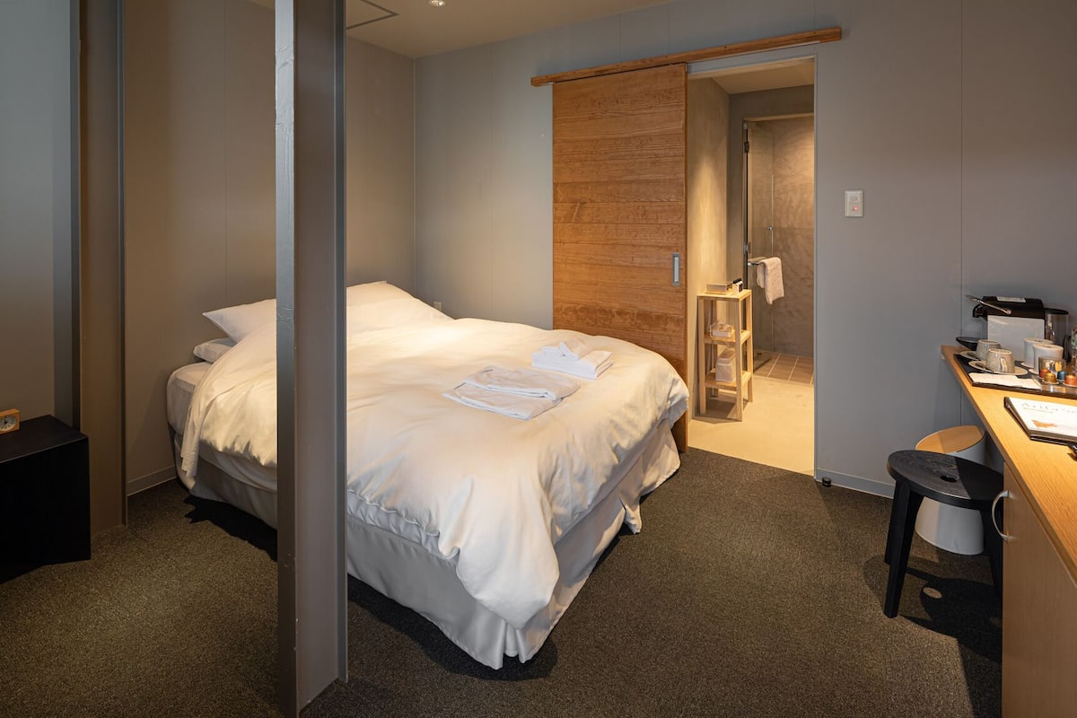 Quarto de hotel com cama, escrivaninha com cadeira, mesa de cabeceira e banheiro visível por uma porta aberta.