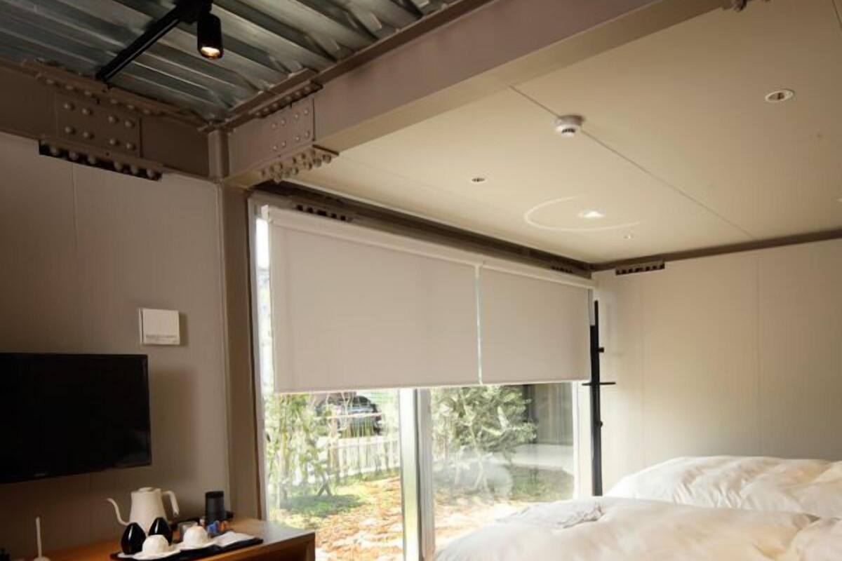 Quarto moderno com uma cama, uma escrivaninha com cadeira, uma televisão e uma janela grande.