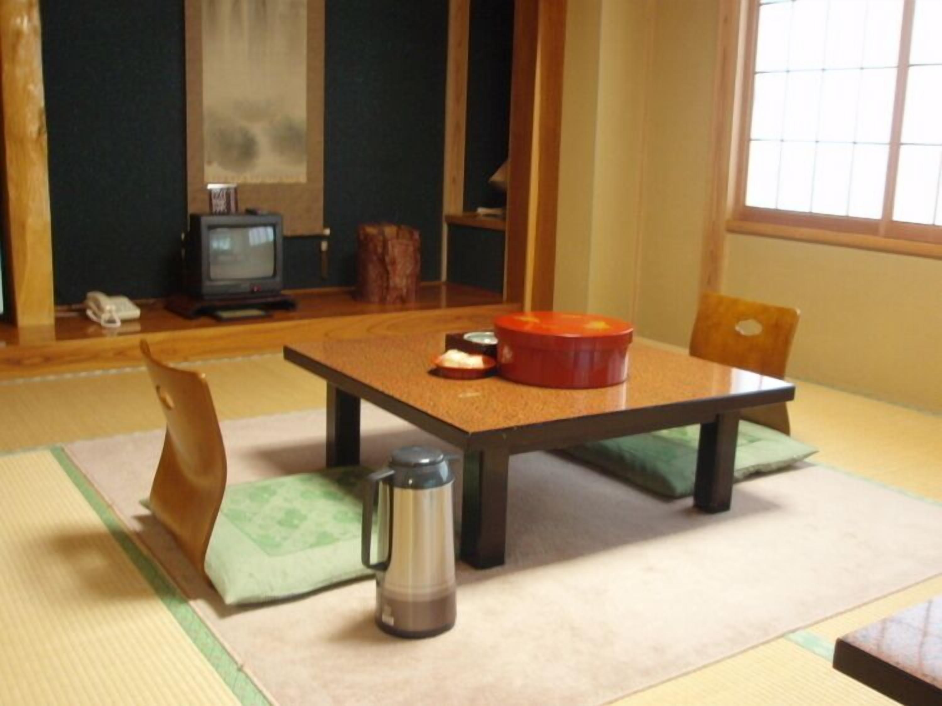 A traditional Japanese room with tatami flooring, a low table with cushions, a small heater, and a television in the background.