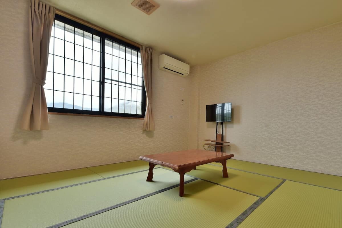 Ein Zimmer mit Tatami-Böden, einer Holzbank, einem Fernseher und einem Fenster mit Vorhängen.