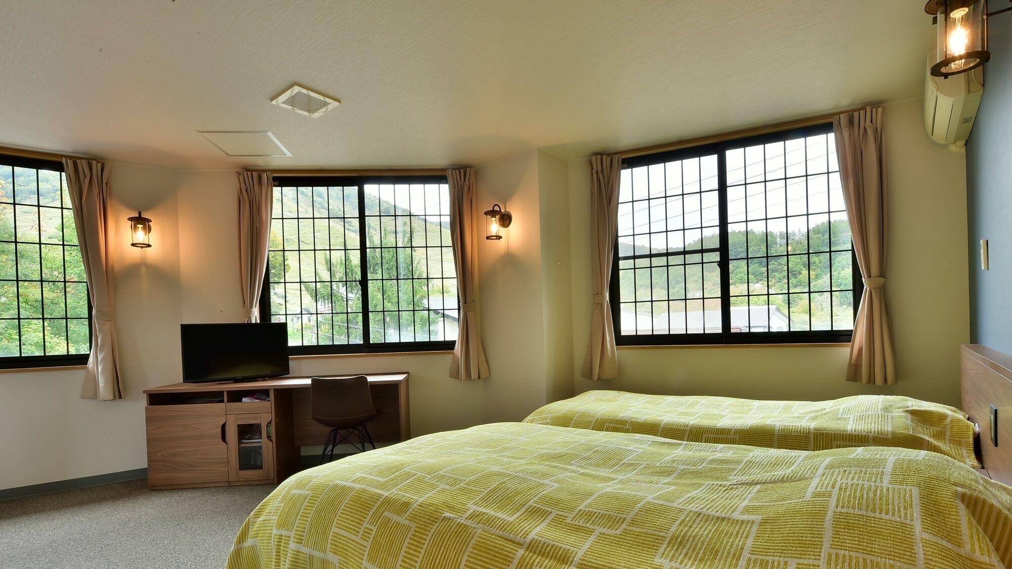 A hotel room with two beds, a desk with a TV, and large windows with curtains.