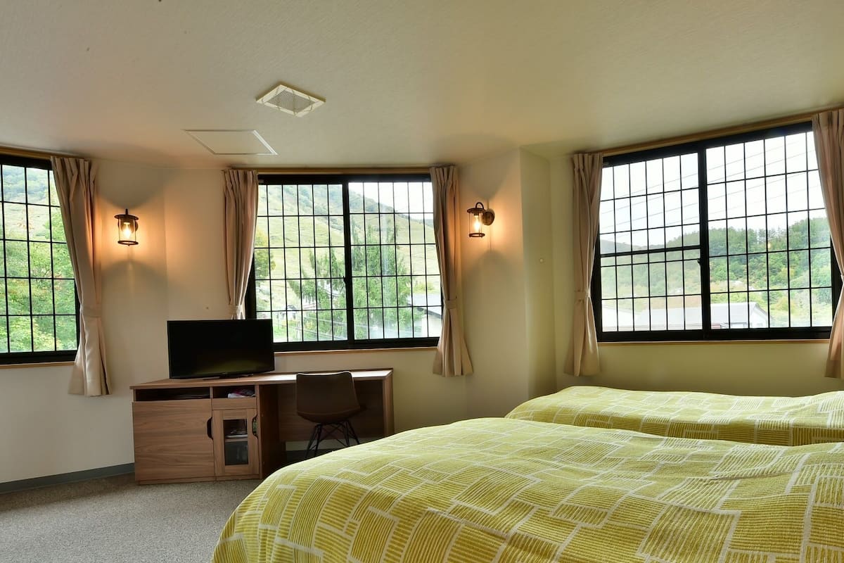 Ein Hotelzimmer mit zwei Betten, einem Schreibtisch mit Fernseher und großen Fenstern mit Vorhängen.