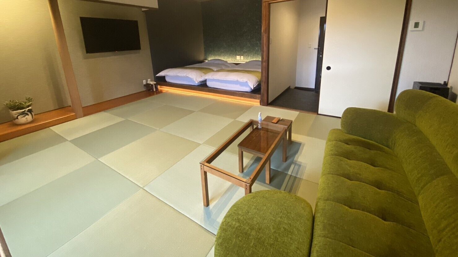 A traditional Japanese-style room with tatami flooring, a low wooden table, a green sofa, and a bed with white bedding.