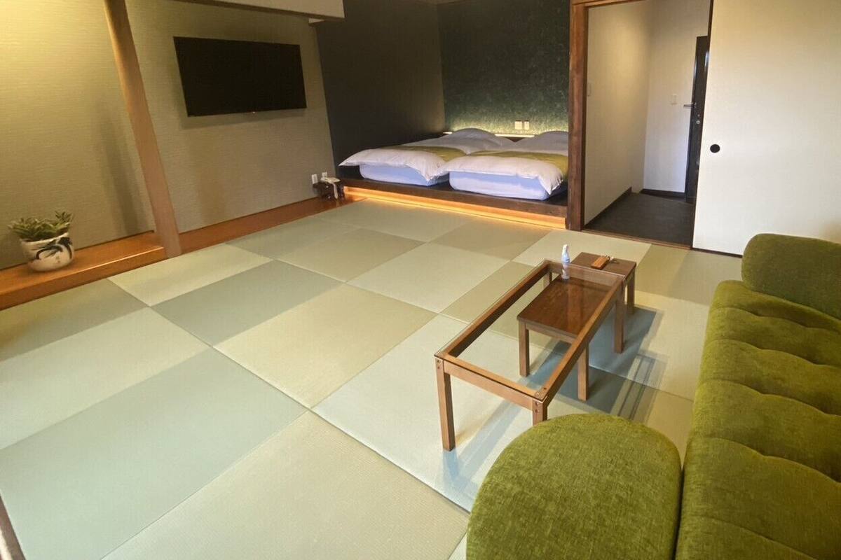 Quarto japonês tradicional com tatames, uma mesa de madeira baixa, um sofá verde e uma cama com roupa de cama branca.