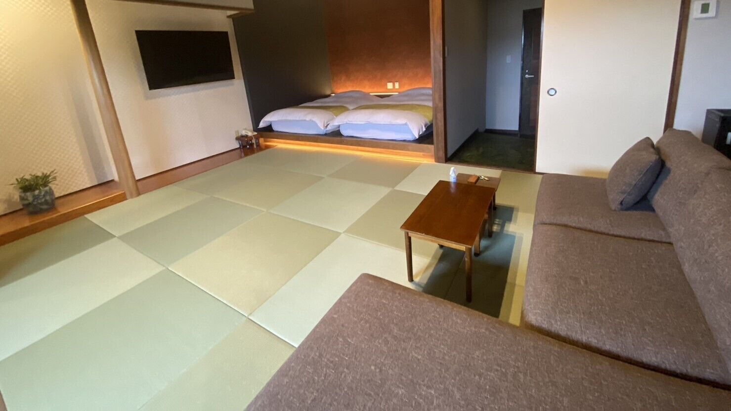 A room with tatami flooring, a sofa, a small wooden table, and a bed with pillows.