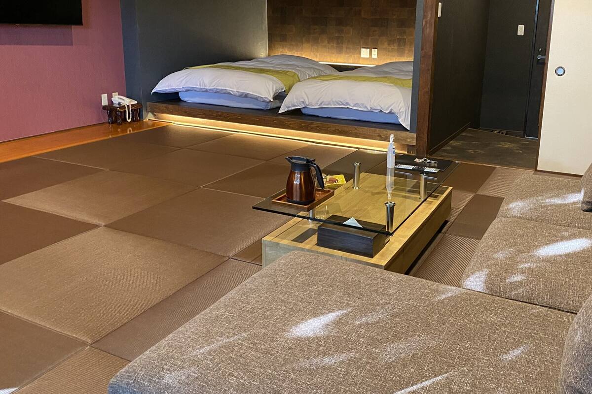 Quarto de hotel moderno com sofá, mesa de centro de vidro, cama com cabeceira e pequena mesa com telefone e chaleira.