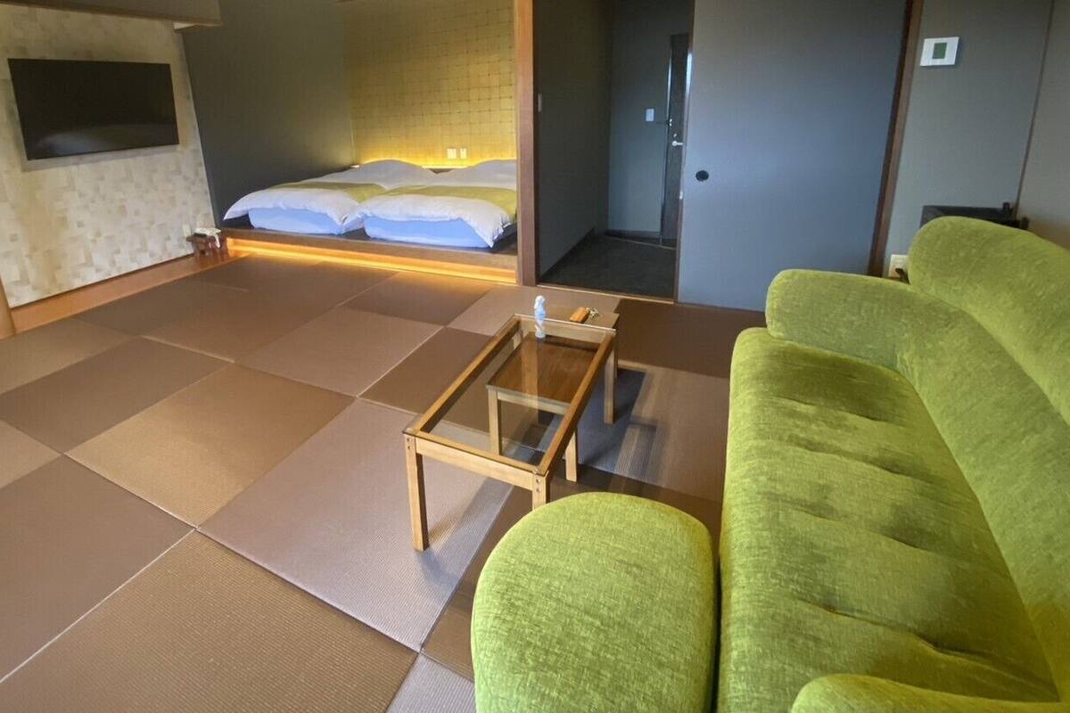 Quarto com piso de tatame, cama com roupa de cama branca, sofá verde e uma pequena mesa de madeira.