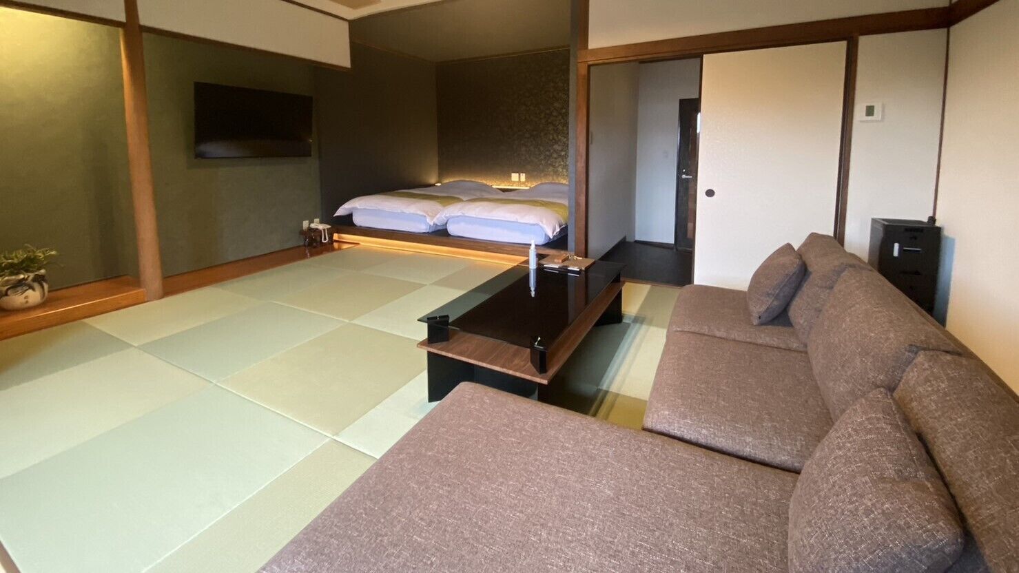A traditional Japanese-style room with tatami flooring, a low wooden bed, a sofa, and a television on the wall.
