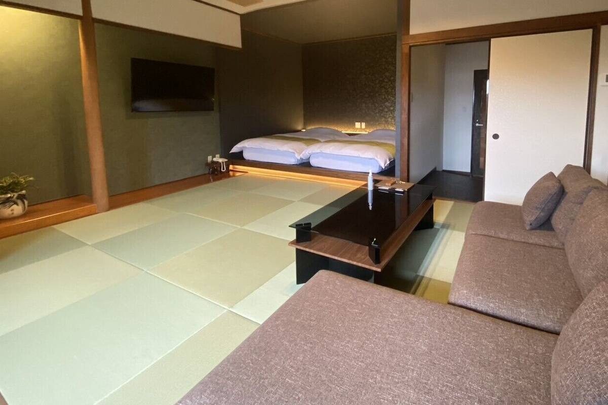 Um quarto tradicional japonês, com tatames, cama de madeira baixa, sofá e televisão na parede.