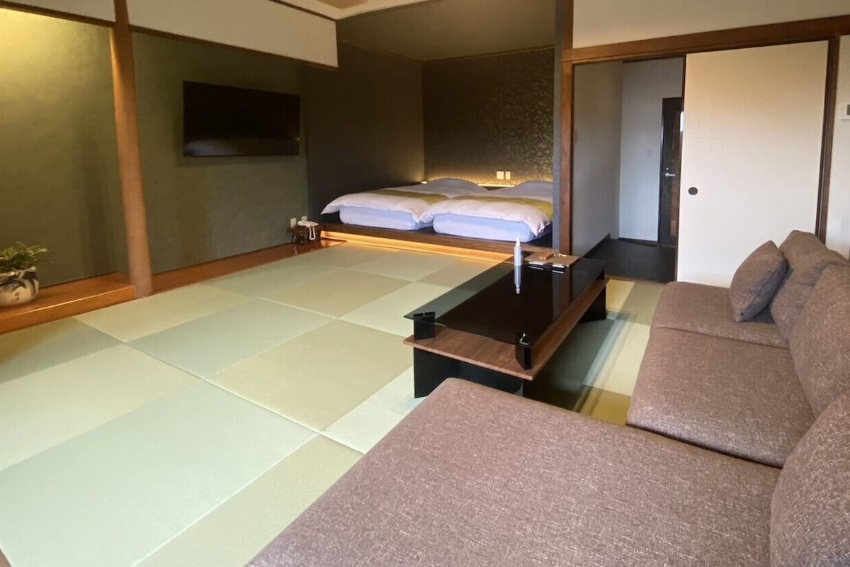 A traditional Japanese-style room with tatami flooring, a low wooden bed, a sofa, and a television on the wall.