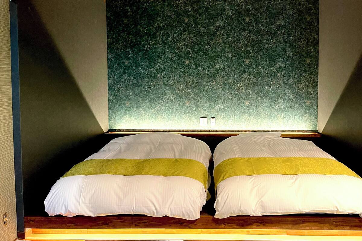 Quarto com duas camas, parede verde com textura e chão de madeira.